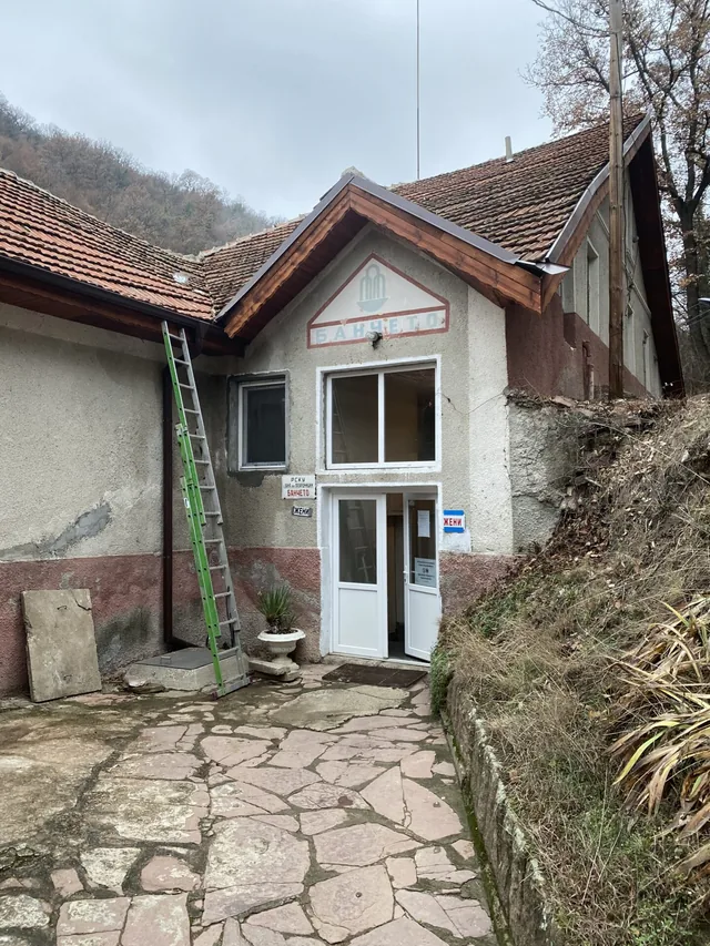 Входът на „Банчето“ — ниската сграда с бялата врата и каменната площадка пред нея.